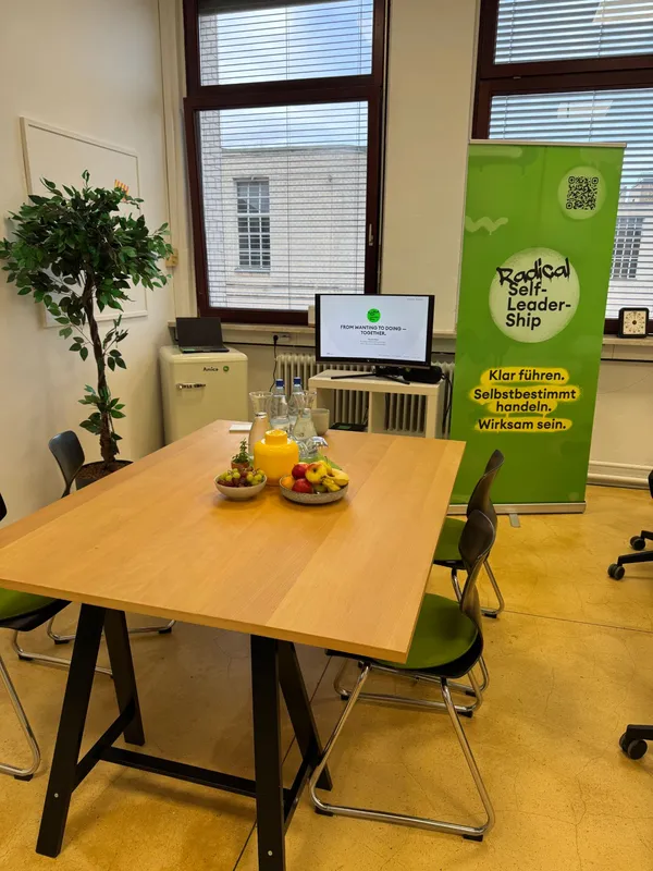 Vorbereiteter Workshop-Raum mit Holztisch, Obst, Wasser, Bildschirm und Radical Self-Leadership Roll-up