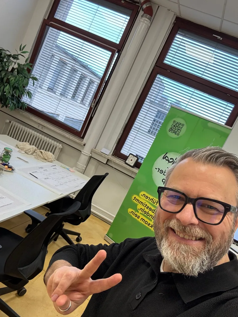 Thorsten Reitz zeigt lächelnd das Peace-Zeichen im Workshop-Raum, hinter ihm das Radical Self-Leadership Roll-up
